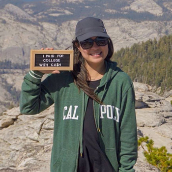 Jaselle stands on top of a mountain wearing a green Cal Poly sweater with a sign that reads “I paid for college with cash.”