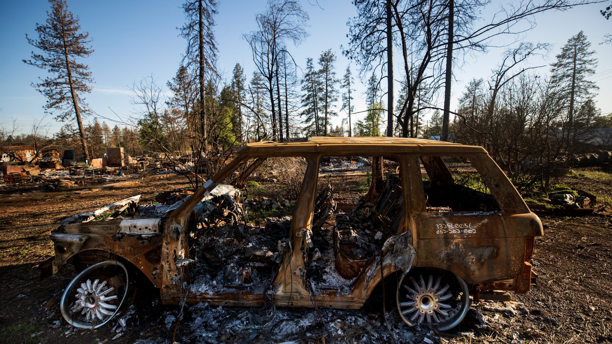 A charred vehicle sits in front of a neighborhood reduced to rubble in Paradise, CA. Photo by Joe Johnston.