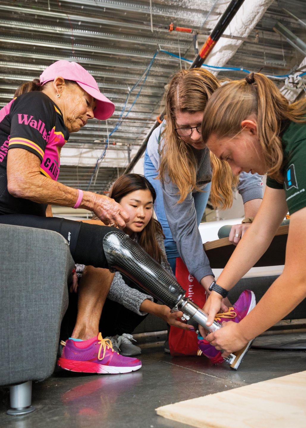 Three female engineering students fit a prosthetic leg onto a local triathlete's right leg in the QL+ lab