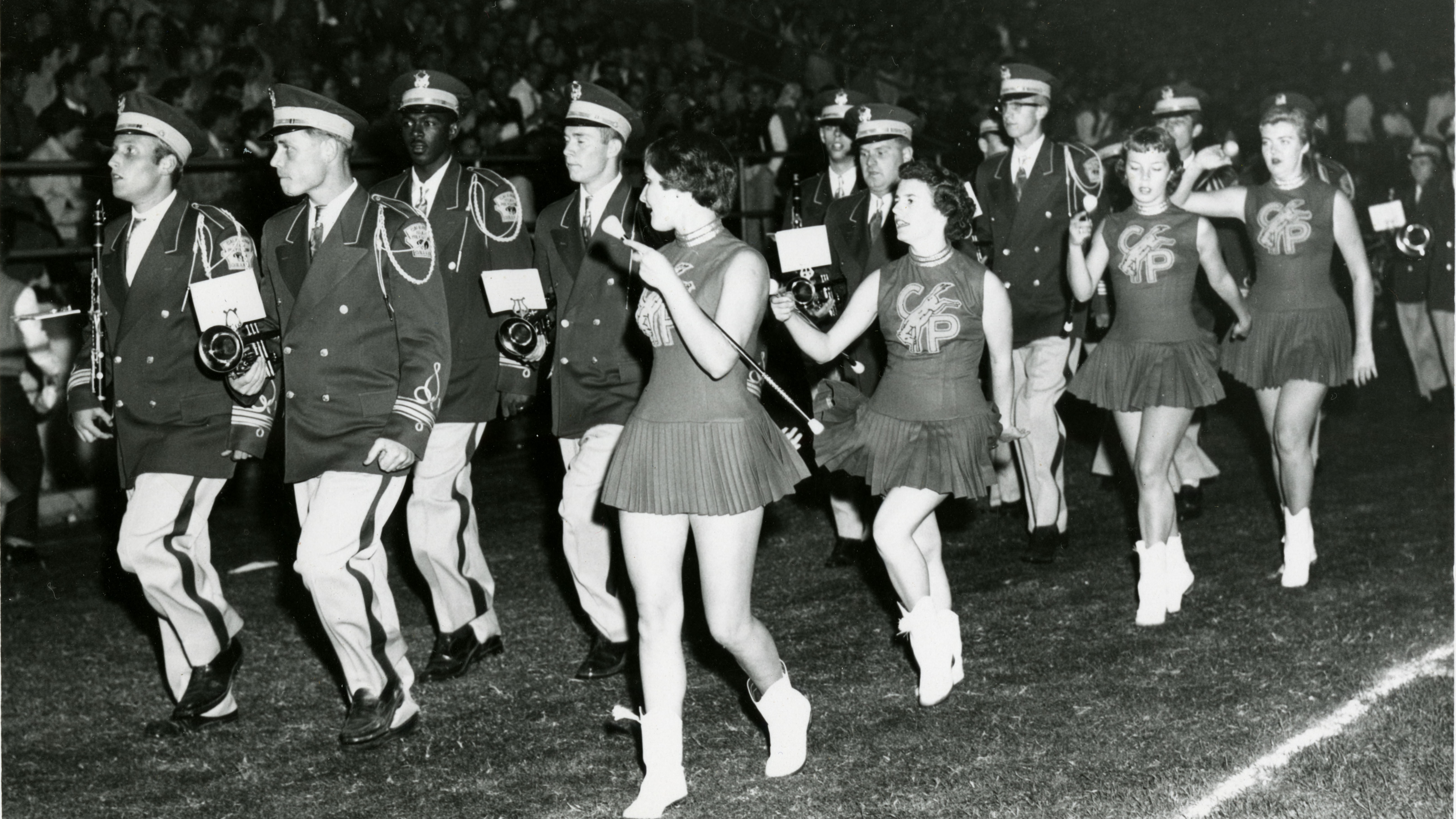 Members of Cal Poly's marching band march alongside female majorettes in a black and white photo