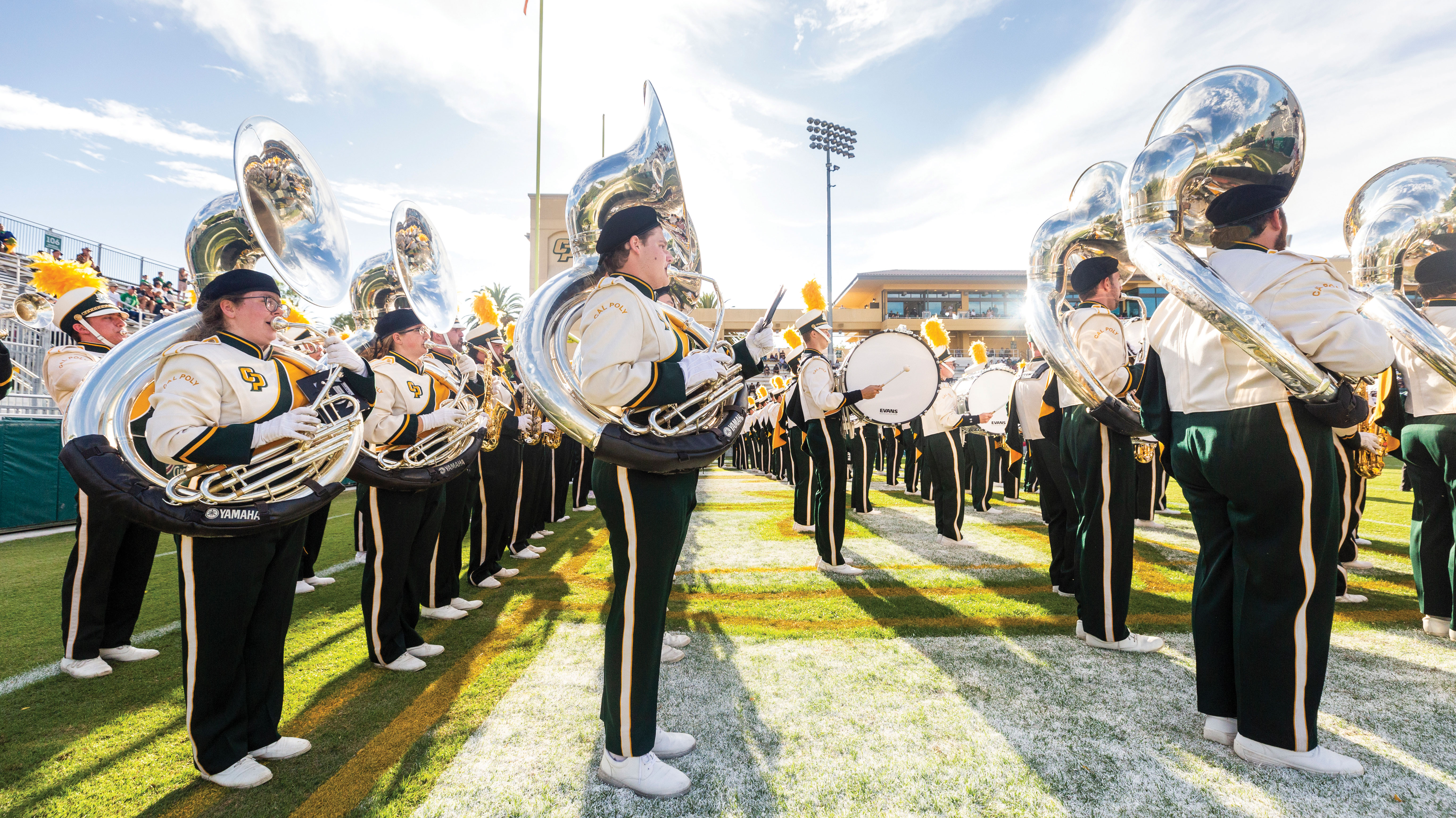 Cal Poly tuba players and drummers stand in formation and play in Spanos Stadium before a football game.