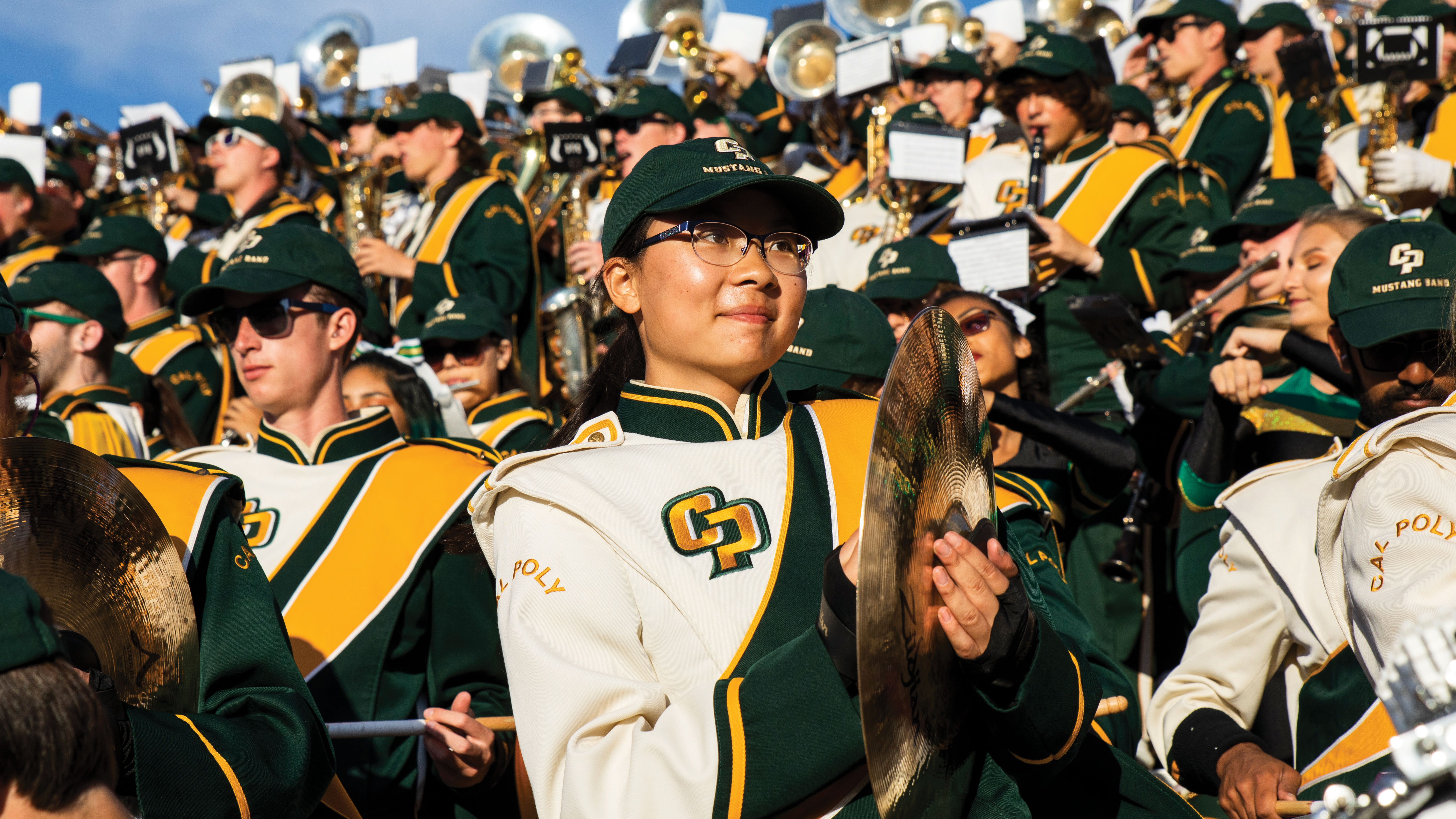 A band member plays the cymbals in the stands of a Cal Poly football game with her fellow musicians behind her.