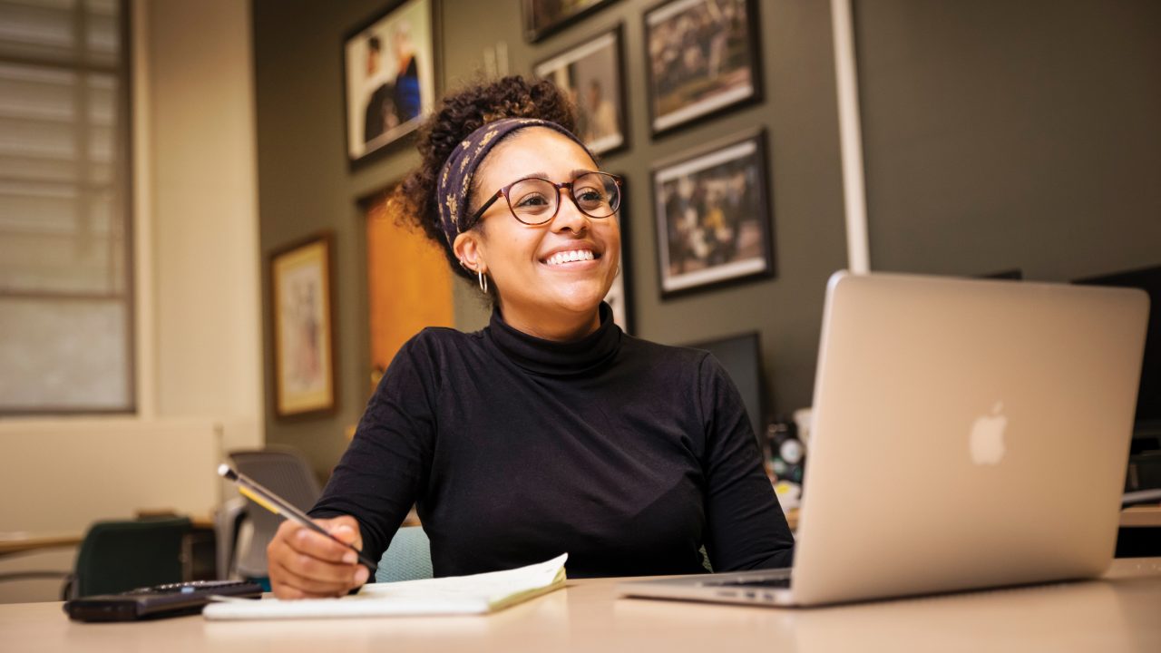 A smiling young woman works in front of a laptop and notebook at Cal Poly's Black Academic Excellence Center.
