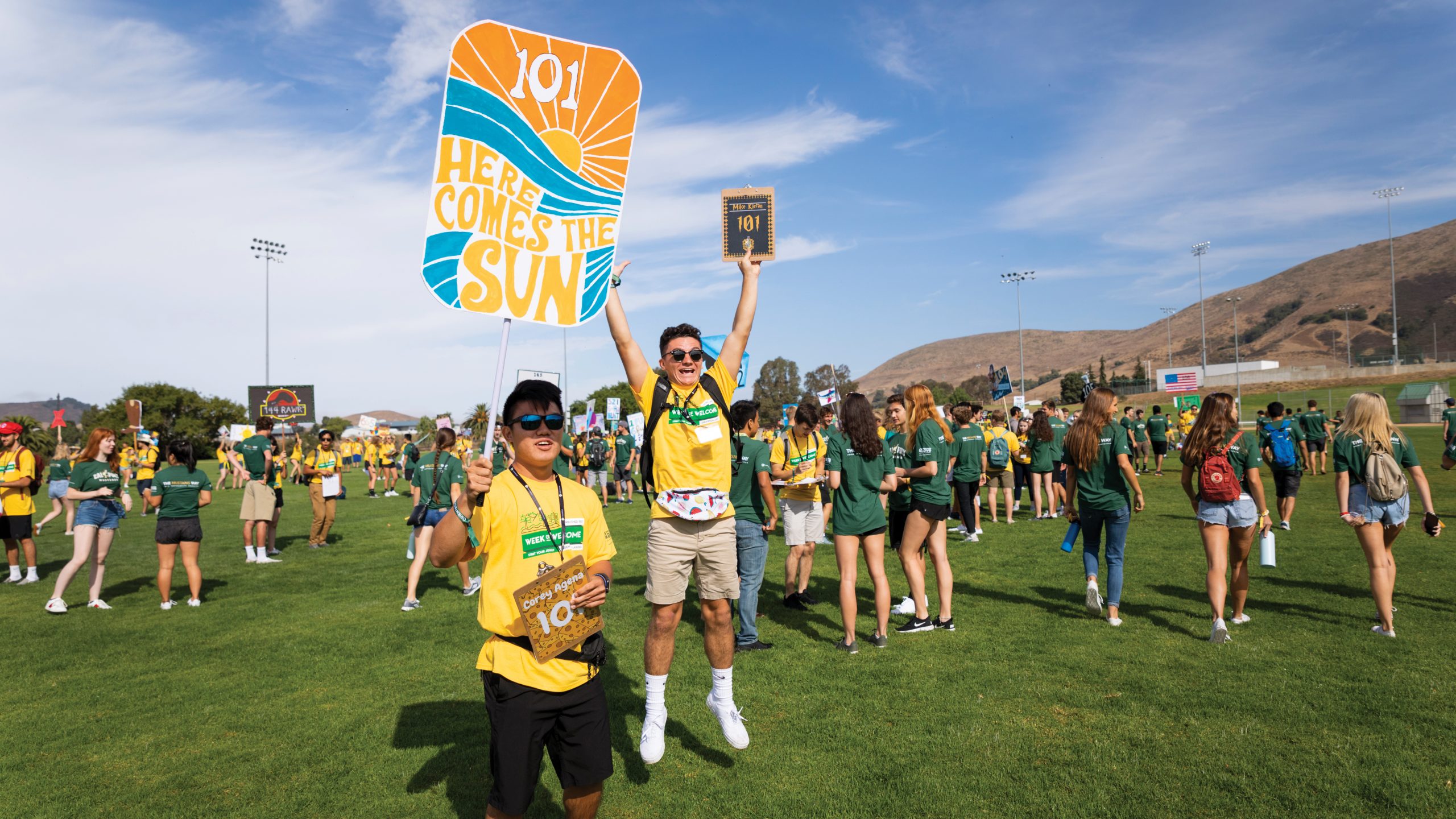 Two young men, wearing sunglasses and yellow Week of Welcome t-shirts and gesturing excitedly with signs, stand on a grassy field in front of dozens of other students.