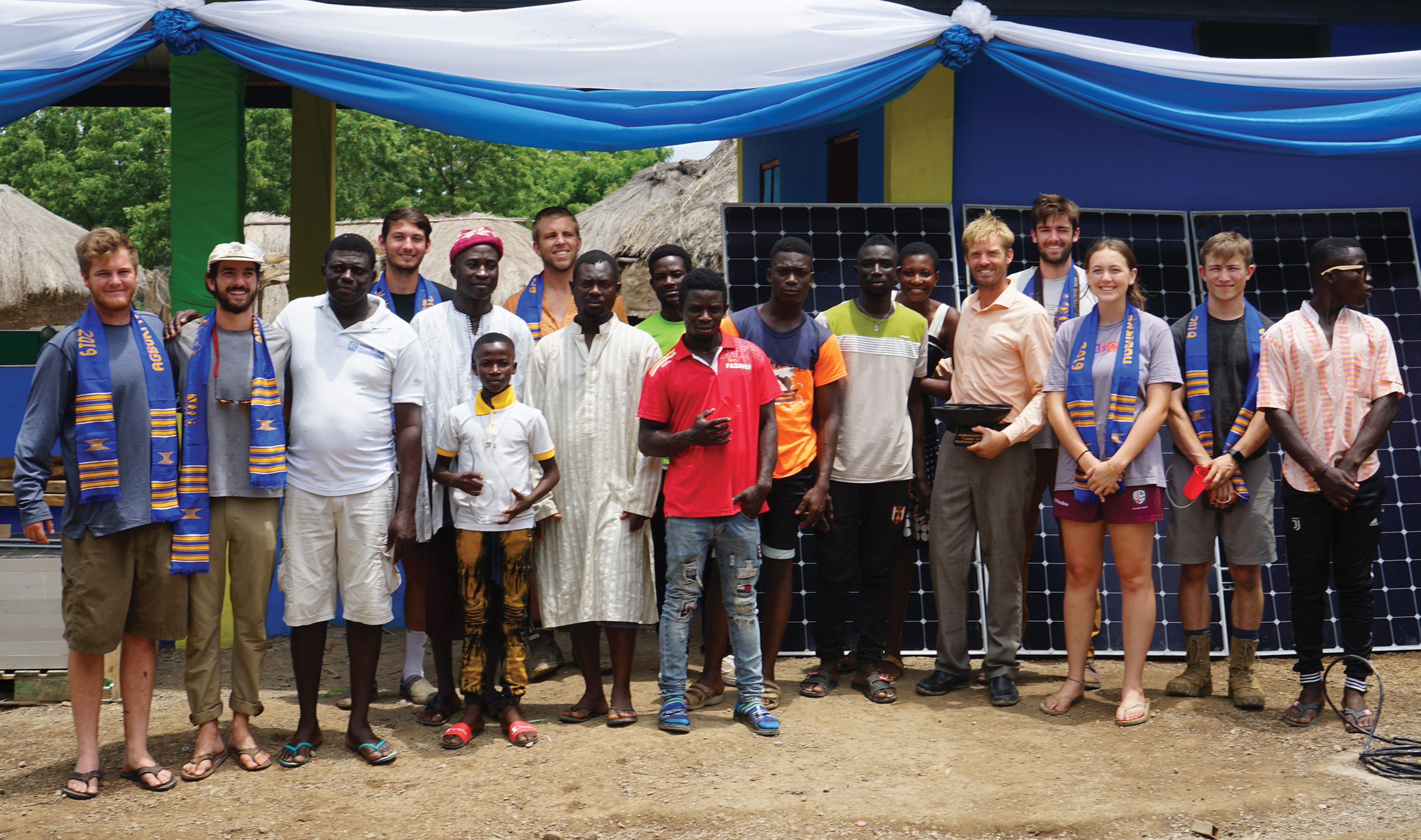 Eight Cal Poly students pose with a group of Ghanaian locals in front of a new building and a set of solar panels.