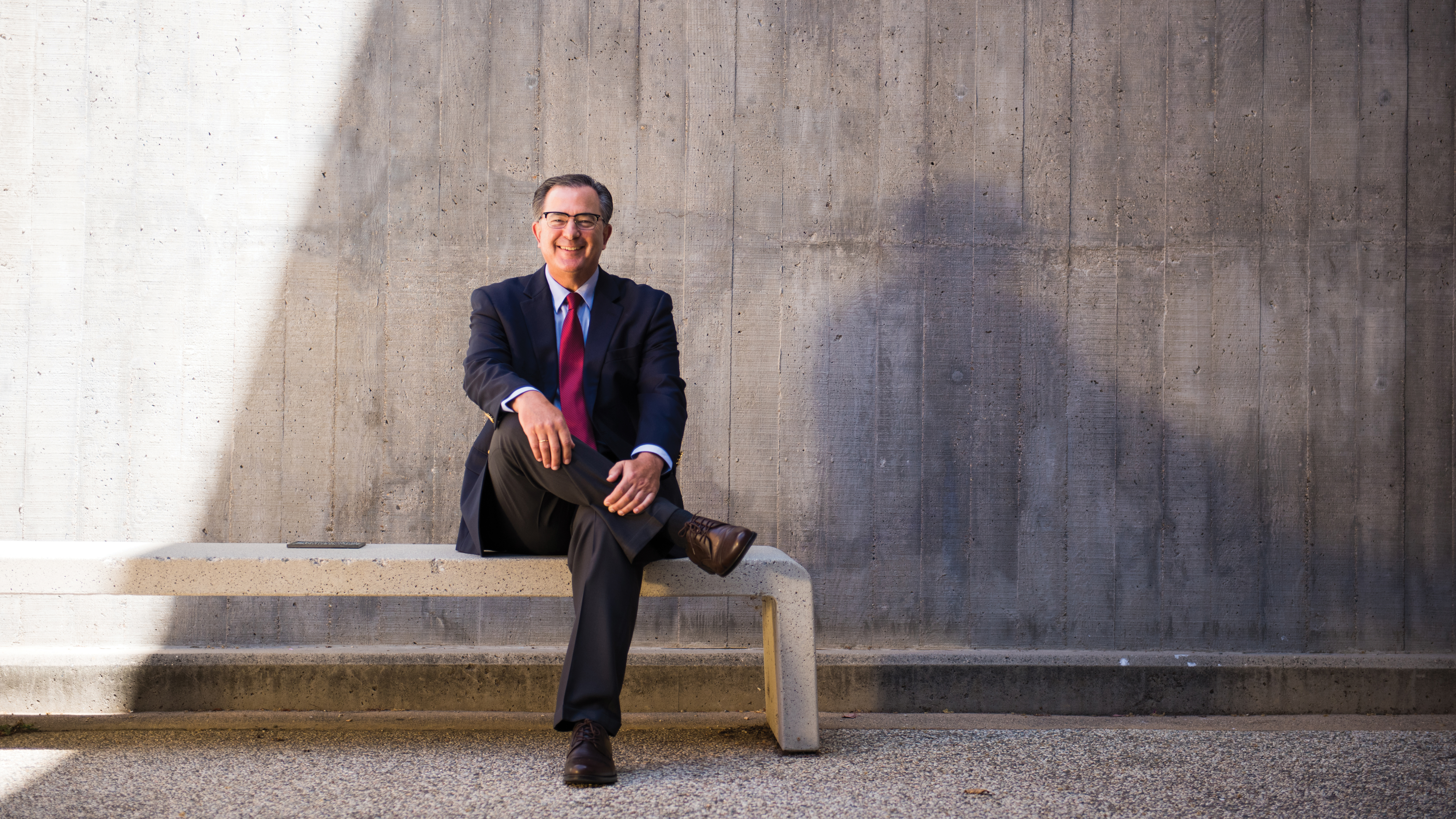 Dean Philip Williams, in a suit and tie, sits on a bench in front of a concrete wall.