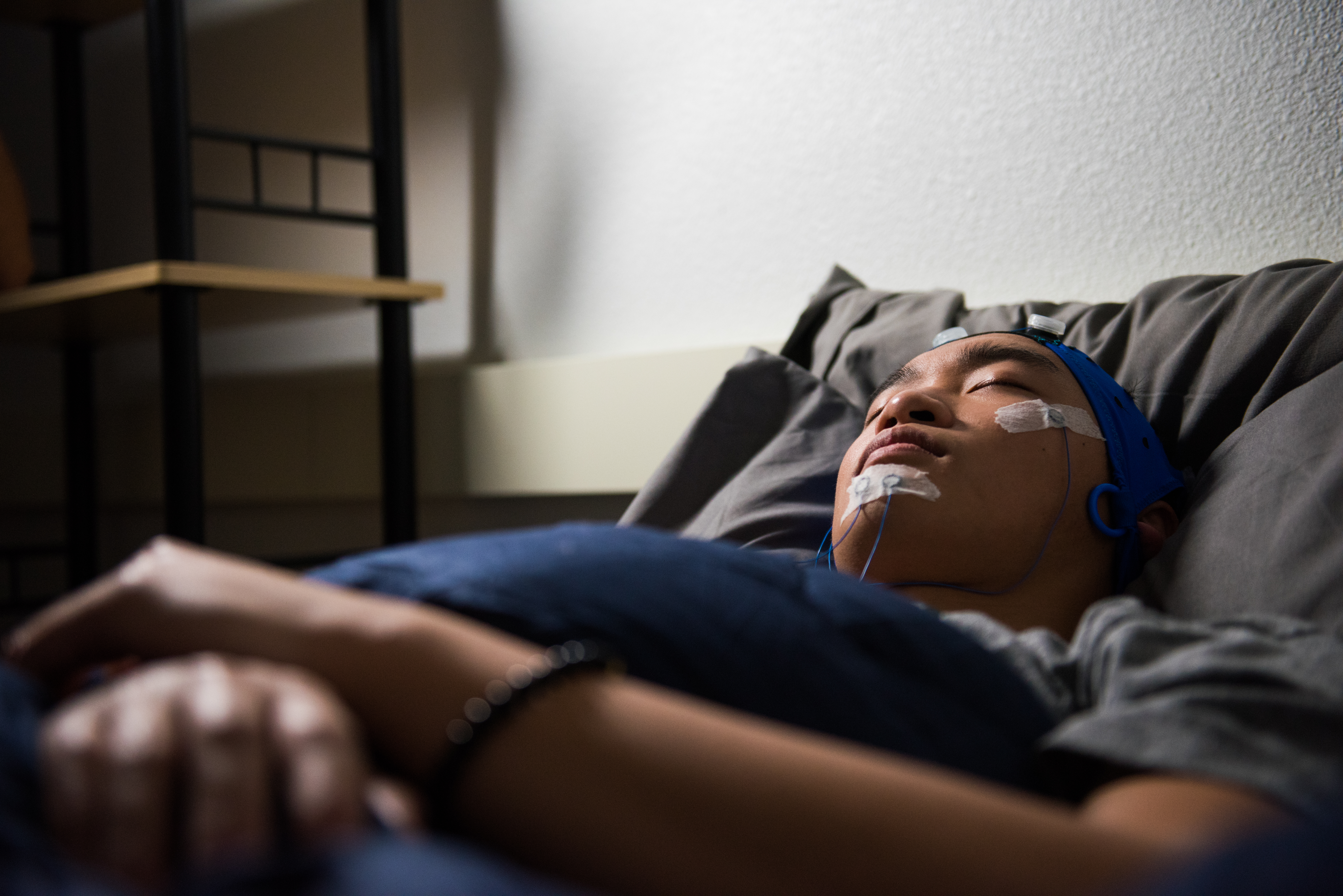 A young man sleeps on a bed in a dimly-lit room, his face connected to several monitoring wires.