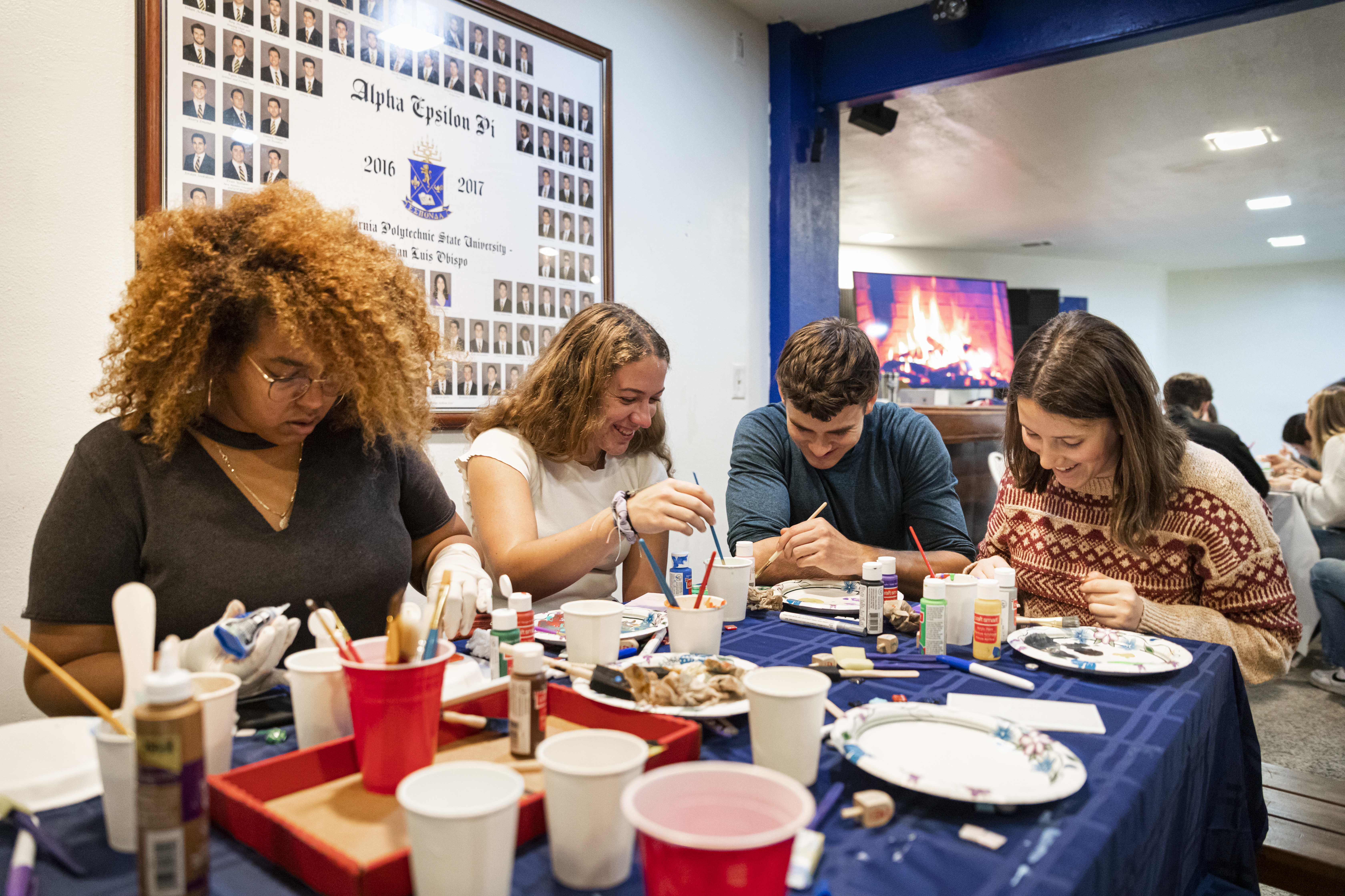 A group of students sit around a table piled with crafting supplies, with a display of fraternity members on the wall behind them and a virtual fireplace burning in the background.