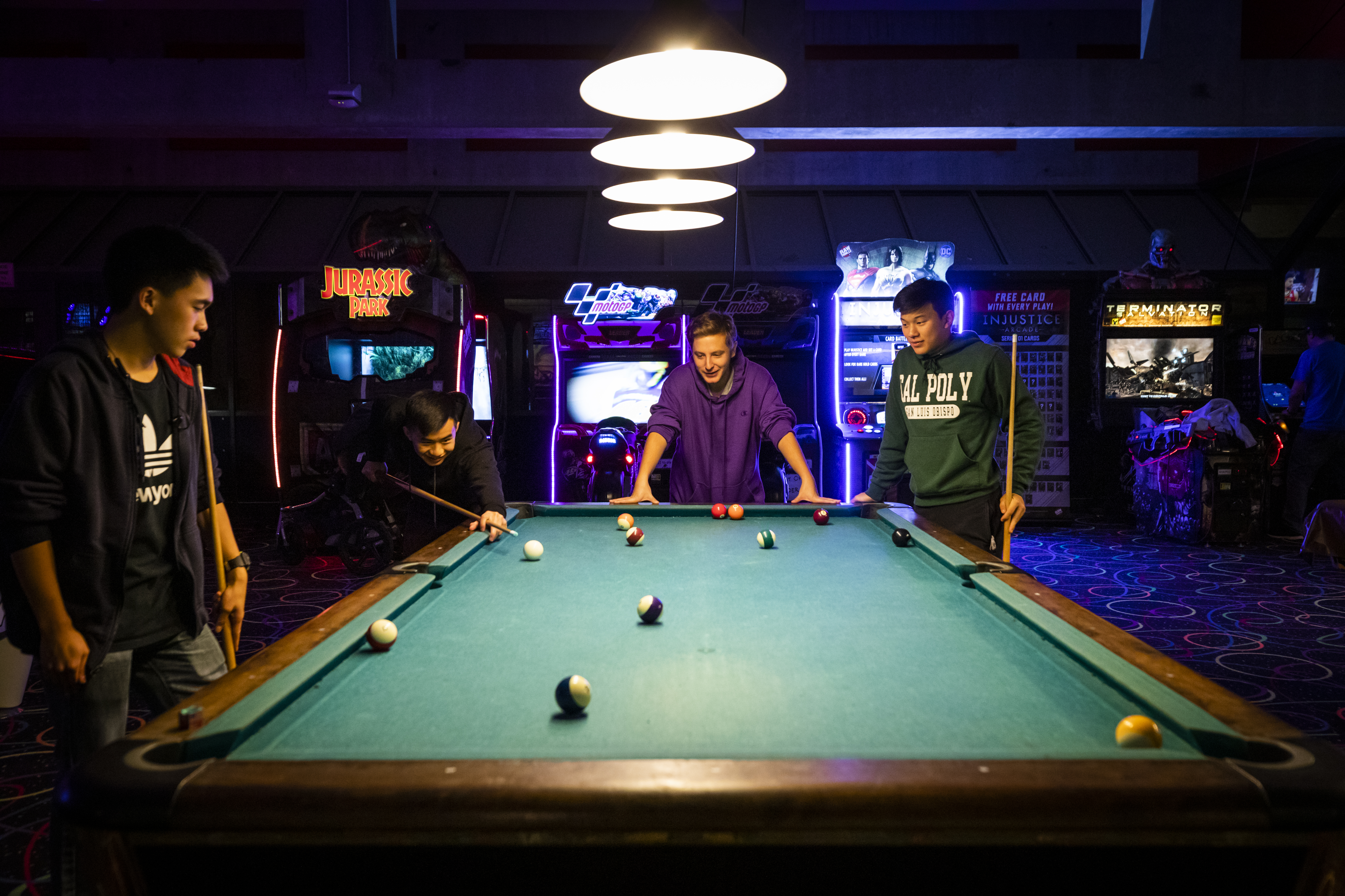 Four young men stand around a pool table in a dark arcade, lit from lamps above and video game cabinets in the backgroun.