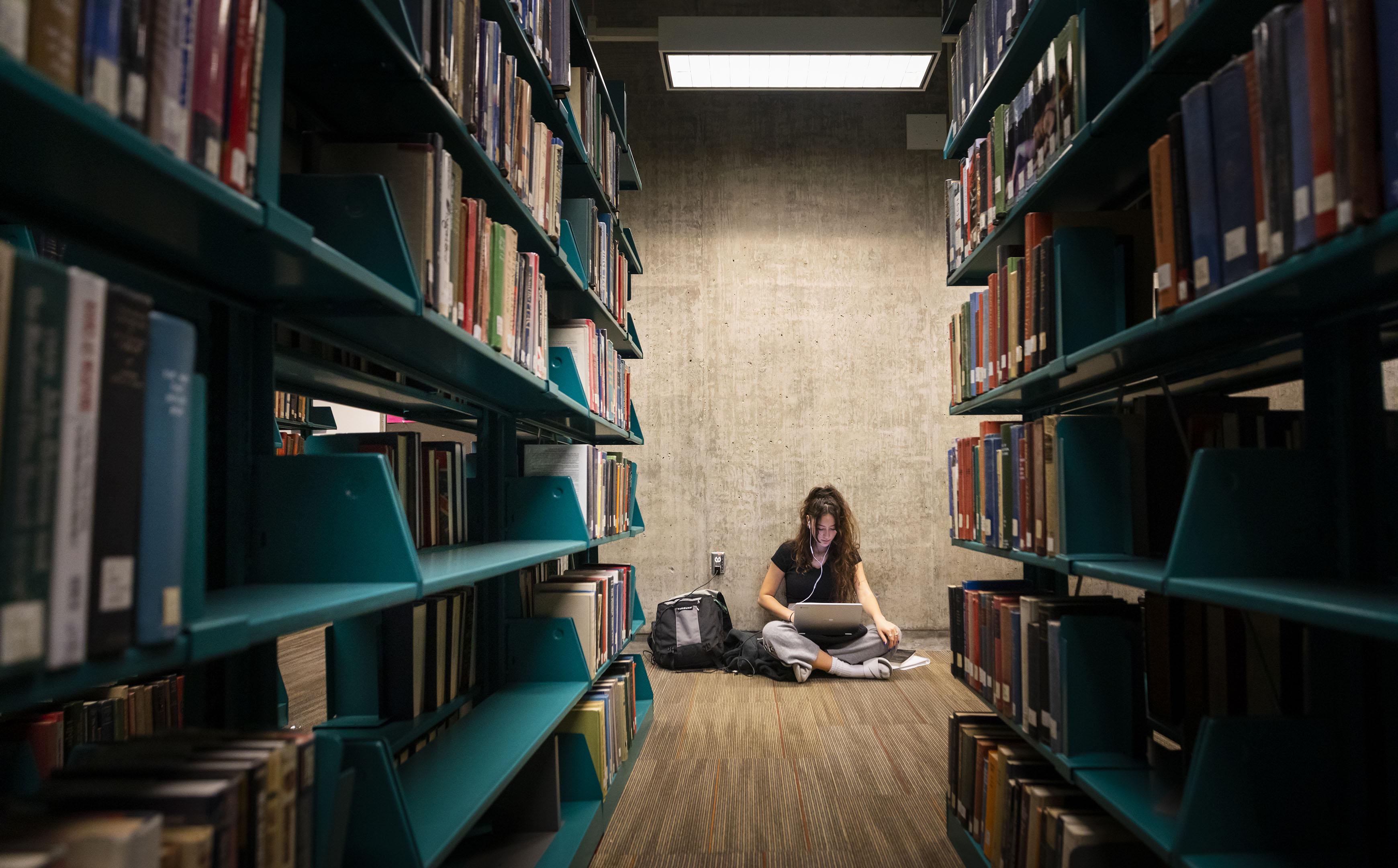 A woman sits on the floor with a laptop and backpack between rows of bookshelves in Cal Poly's Kennedy Library