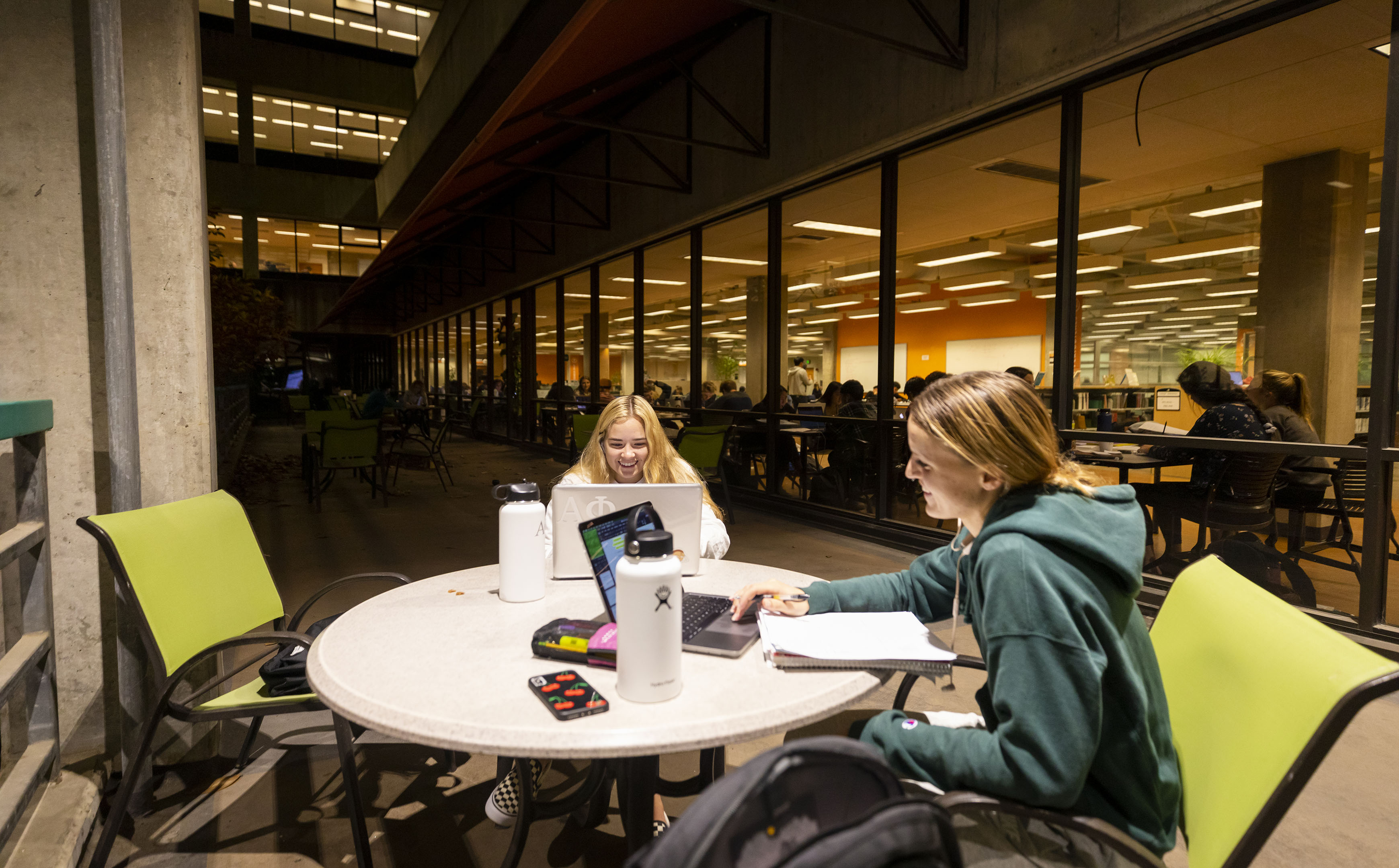 Two female students sit at a table with notes, waterbottles and laptops in Kennedy Library's atrium