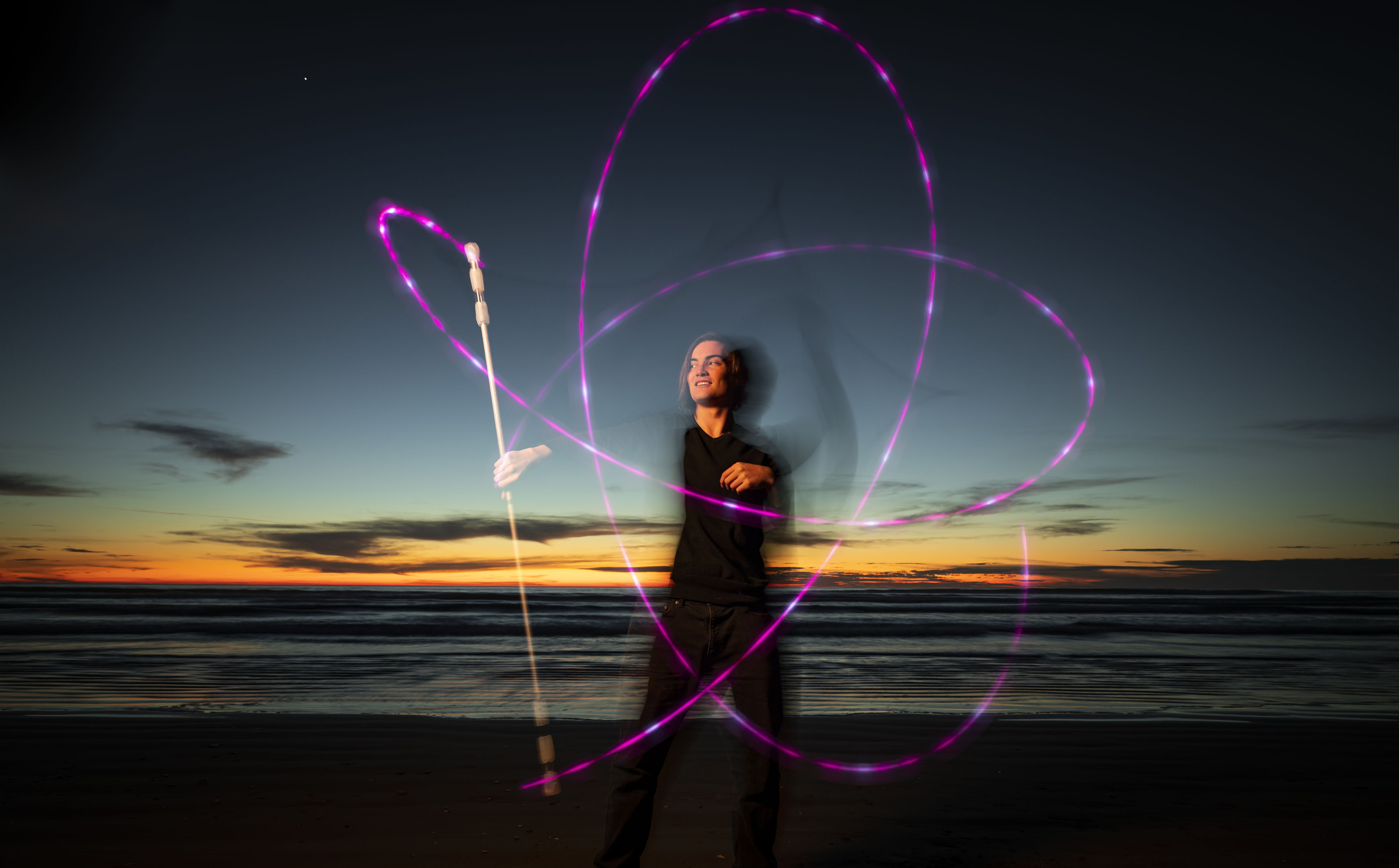 A young man stands on the beach tracing a pattern with a purple light on a long stick as the sun sets in the background