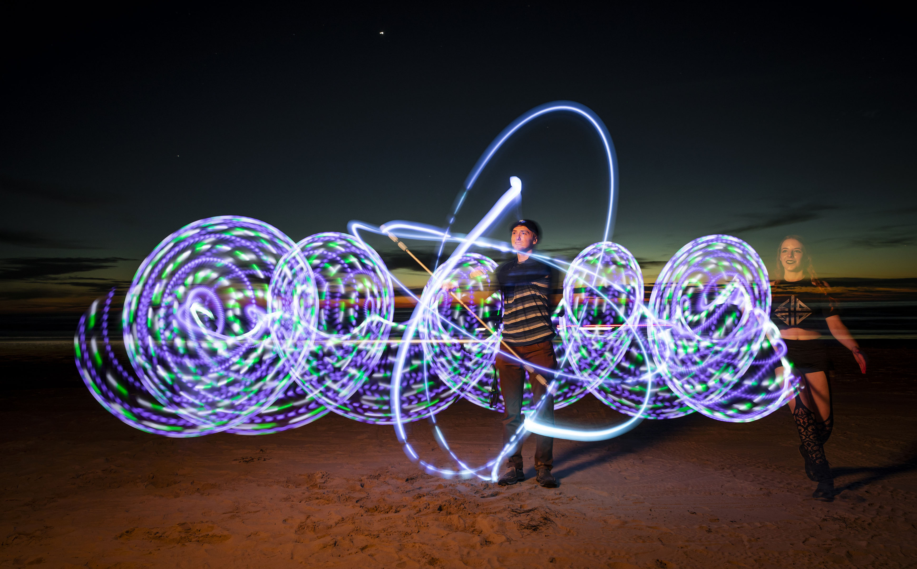 A young woman and man stand on the beach swirling lights to create a purple and green pattern