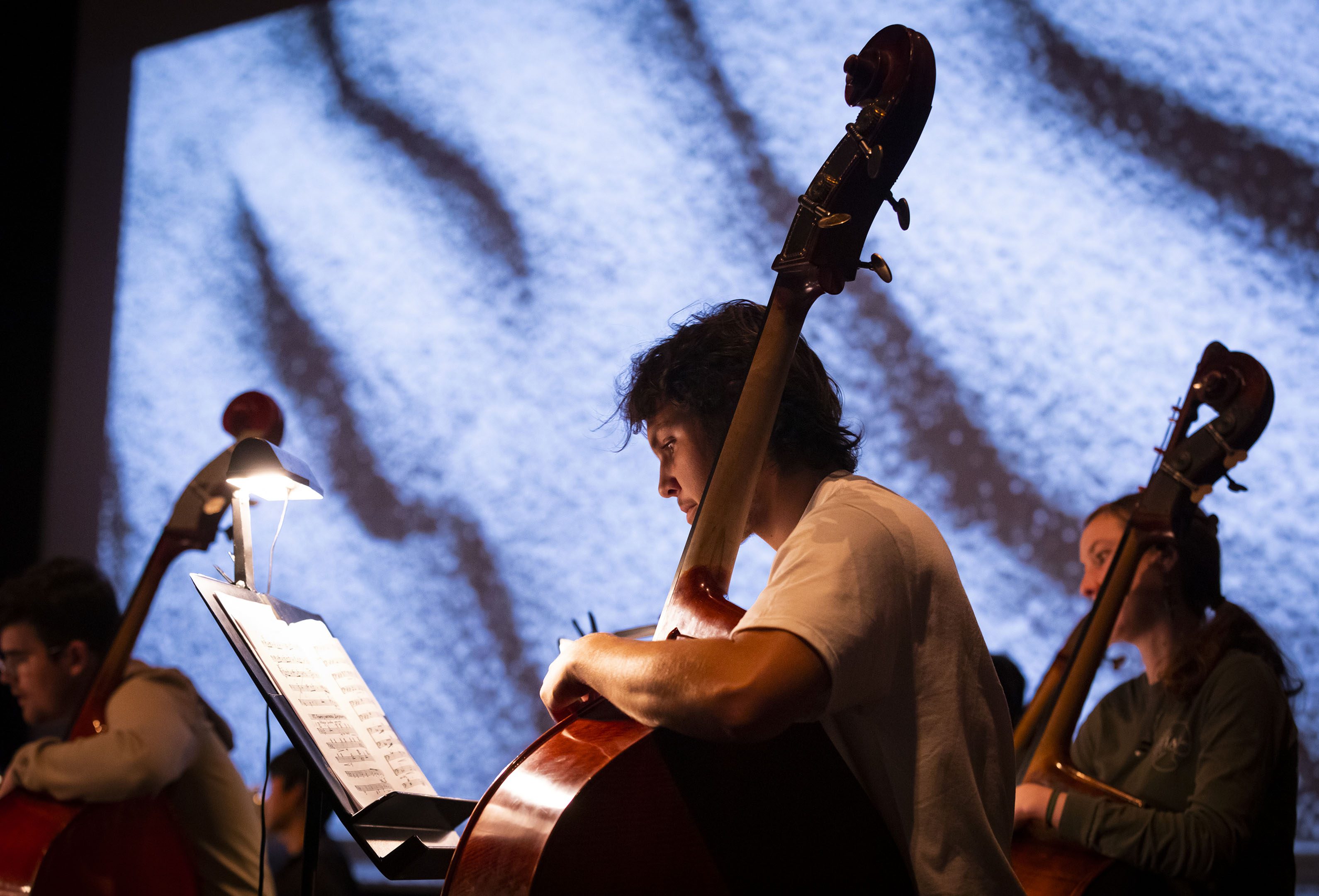 Three musicans play the cello in front of an illuminated screen and sheet music