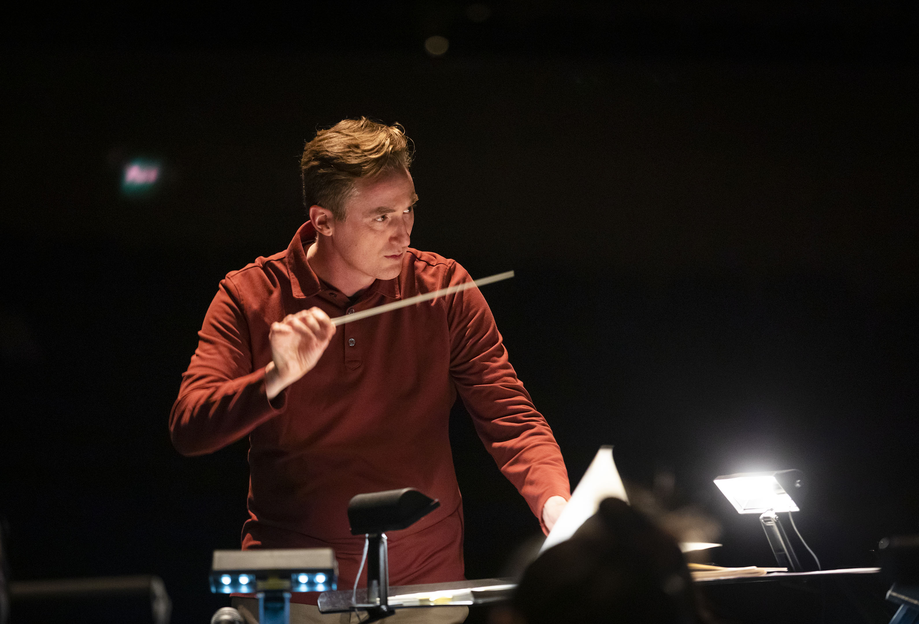 A man in a red shirt conducts musicians with a baton