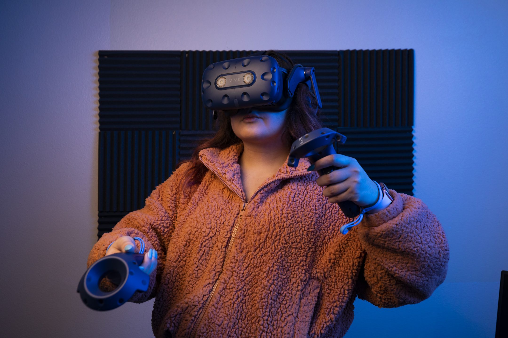 A young woman in an orange sweater and a VR visor handles a set of VR controls.