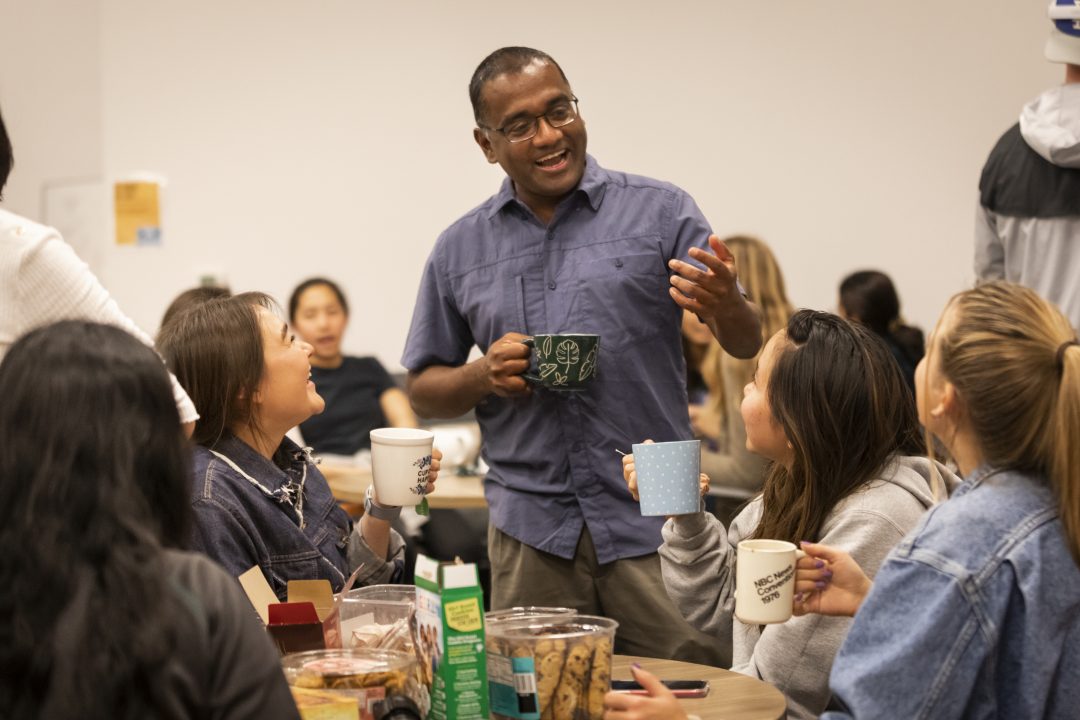 A professor in a blue shirt stands with a tea cup, chatting with students sitting at a table.
