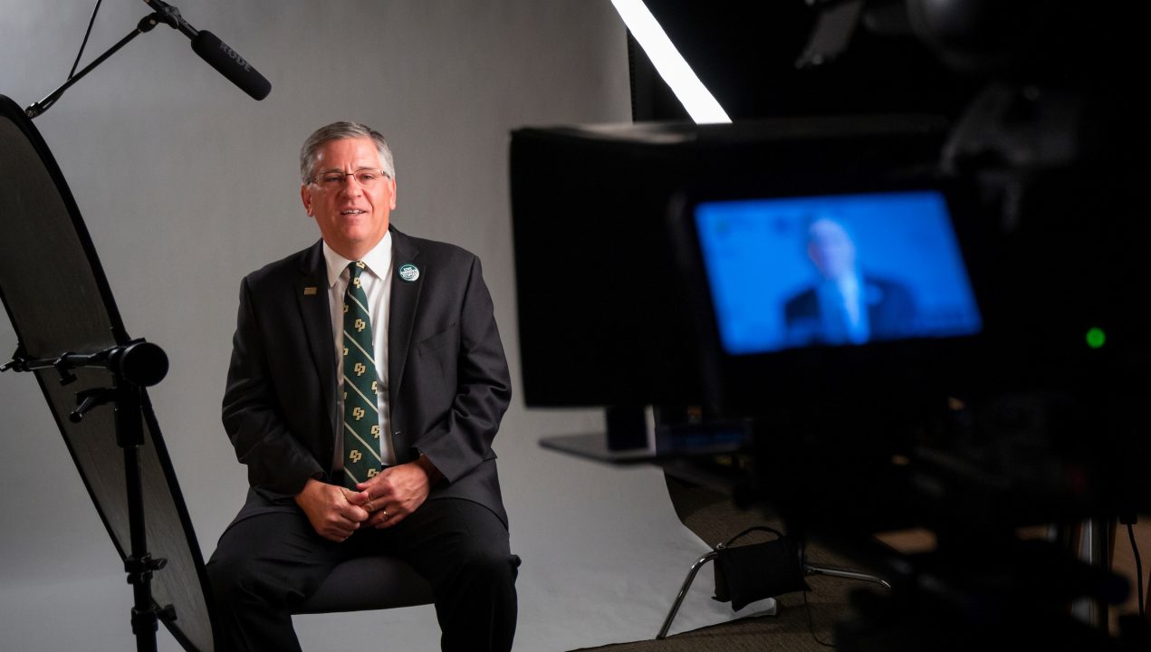 President Armstrong, in a suit and Cal Poly tie, speaks into a video camera and microphone in front of a white backdrop