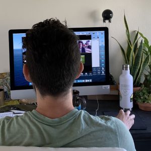 Seen from behind, a man in a green T-shirt faces a computer showing a Zoom meeting.