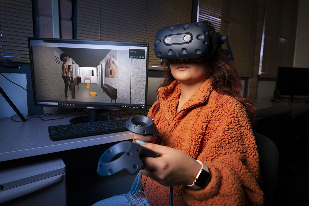 A young woman in an orange sweater pilots a VR experience, in front of a monitor showing a digital rendering of a massage parlor.