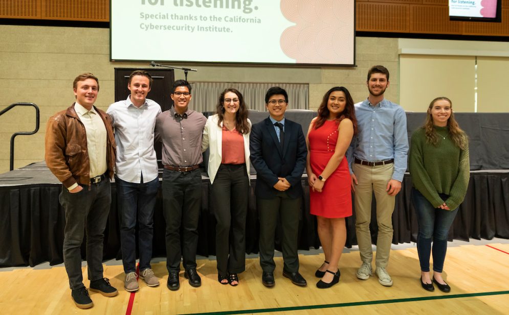Eight students stand in front of a stage in a Cal Poly auditorium.