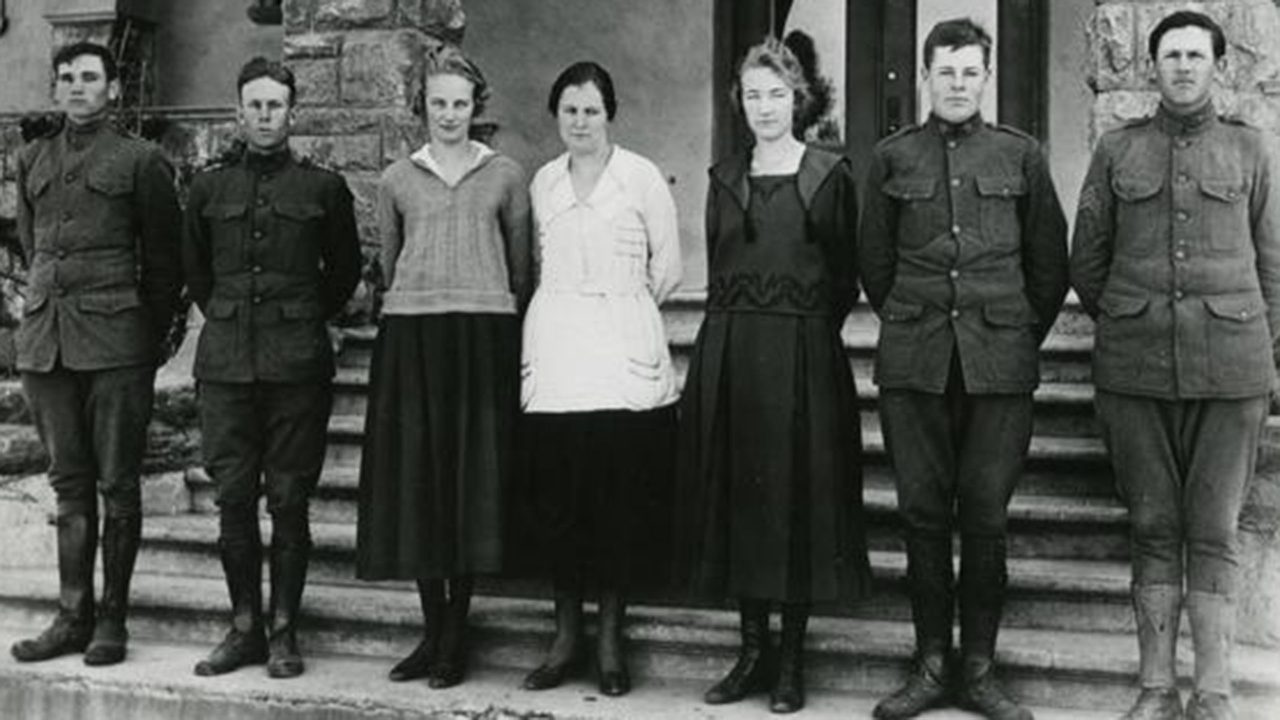 Seven students, men in early 20th-century military uniforms and women in long blouses and skirts, pose in front of a campus building in a vintage photo