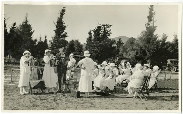 A group of musicians, women in long white dresses and hats, perform instrumental music on a campus lawn in a black and white photo.