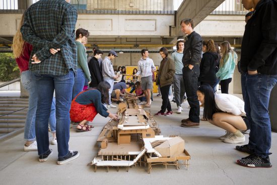 Students gather around wood architectural models displayed on the concrete courtyard of the Architecture building.