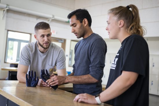Three students gather around a table, examining a prototype prosthetic hand.
