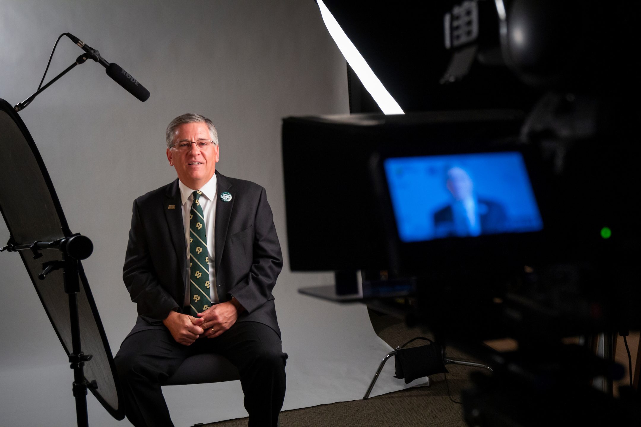 President Armstrong, in a suit and Cal Poly tie, speaks into a video camera and microphone in front of a white backdrop