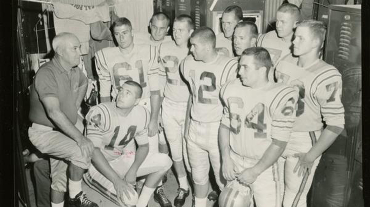 A black and white photo of a football coach and 10 uniformed players in a locker room