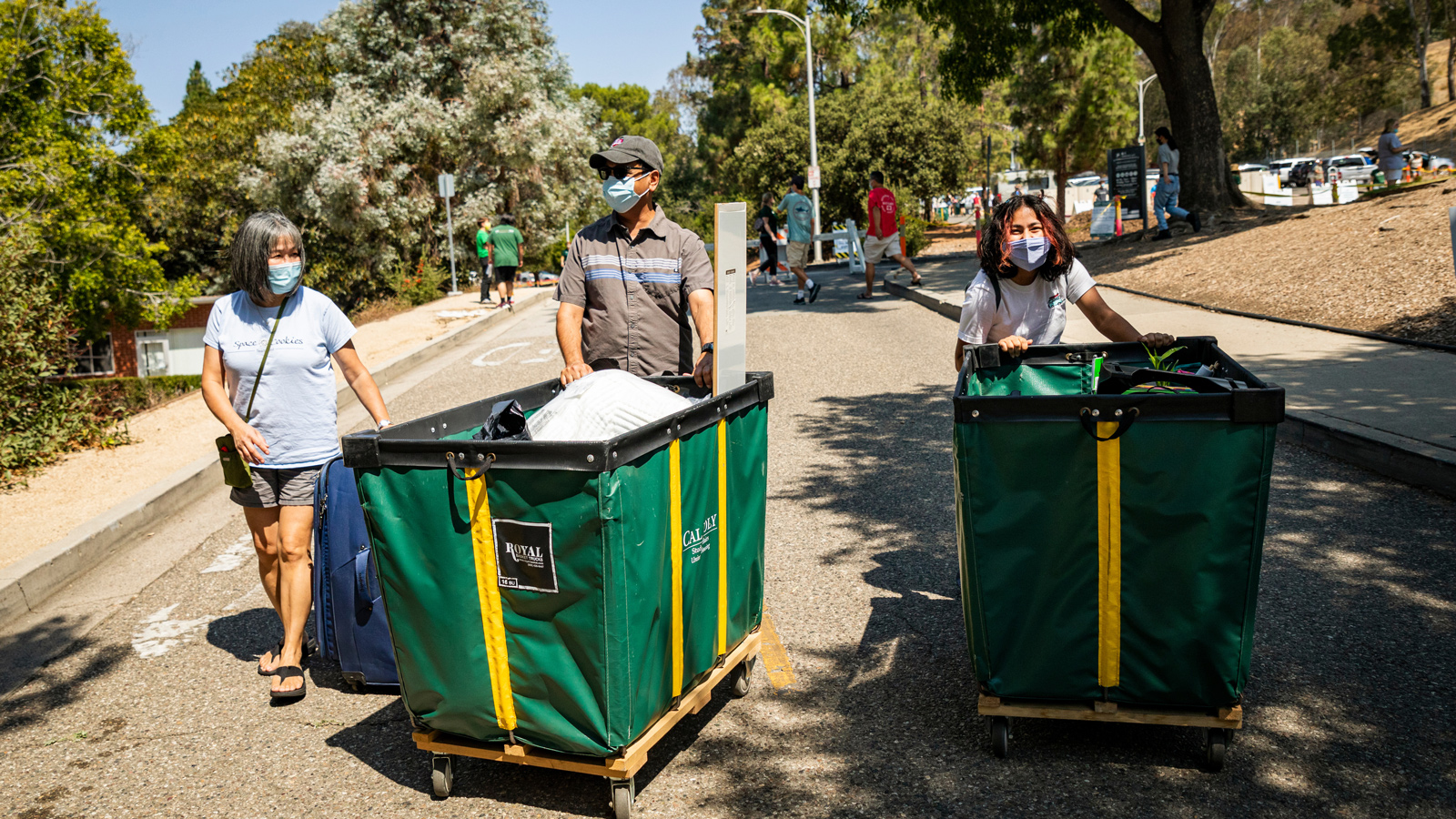 A student and her parents, smiles evident under face coverings, push green and yellow carts loaded with dorm supplies up a campus driveway.