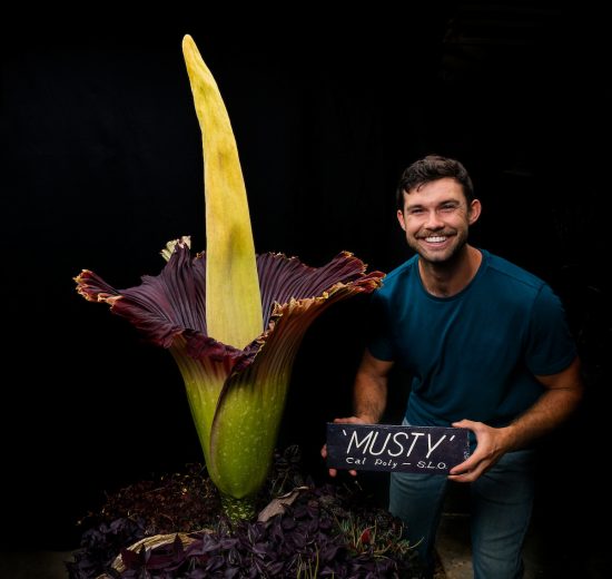 A young man holding a sign reading "Musty" smiles next to a massive tropical corpse flower