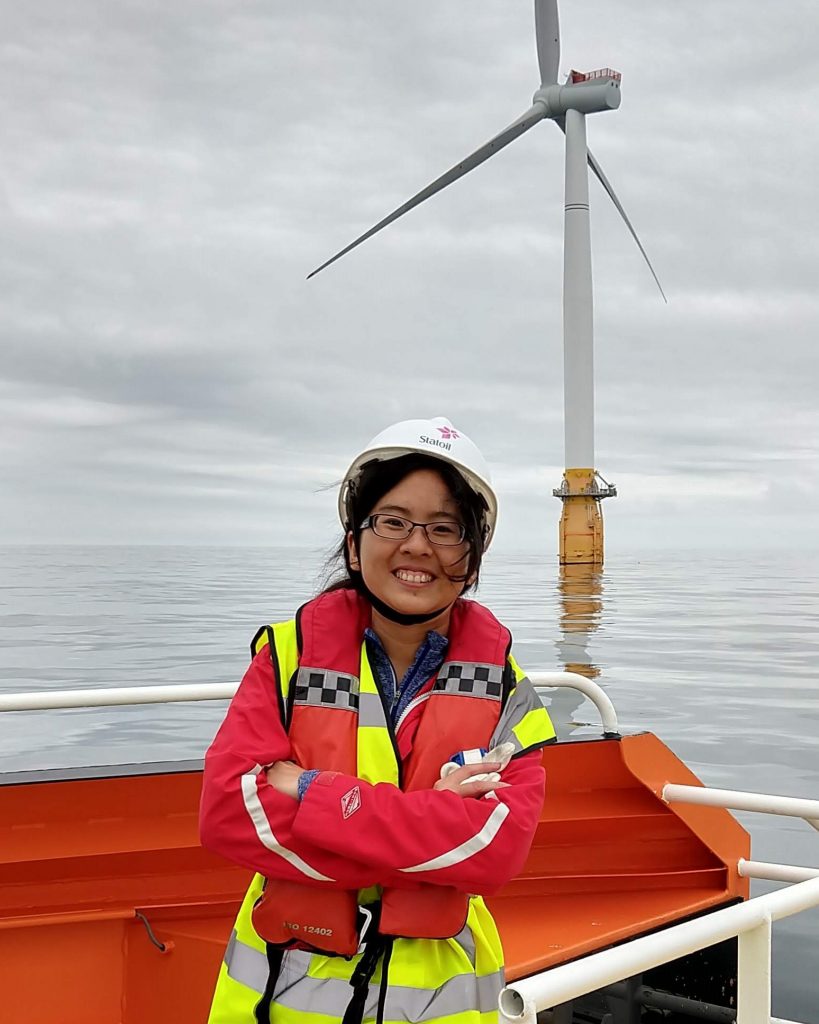 A young woman in a life vest and hardhat smiles on the deck of a boat in front of an ocean-mounted windmill.