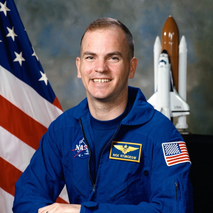 Astronaut Frederick "CJ" Sturckow smiles in his blue flight suit in front of an American flag and a model of the Space Shuttle