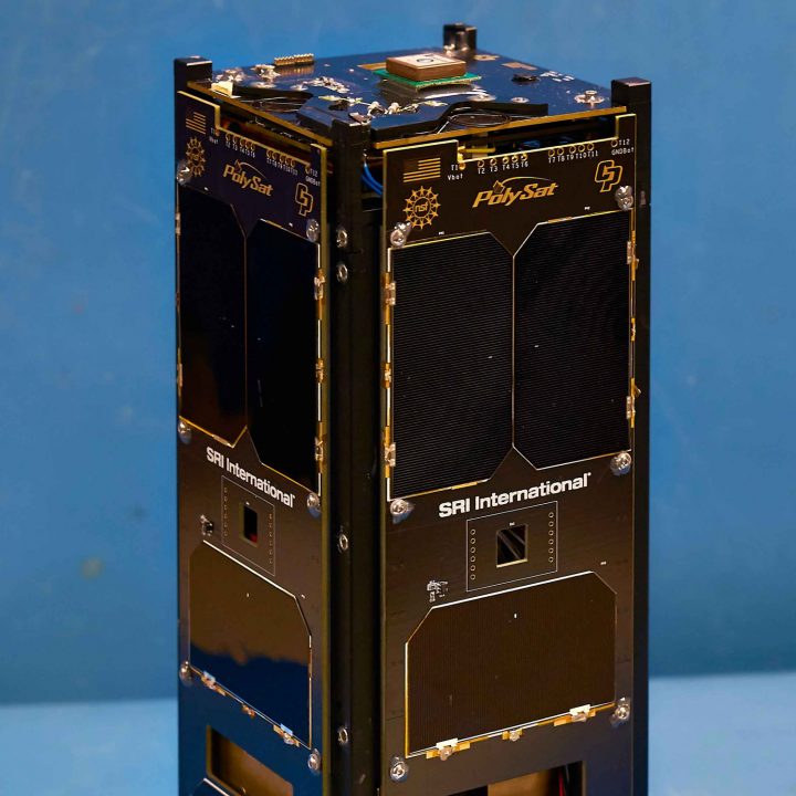 A cubesat with the words 'PolySat' engraved on it stands in front of a blue background