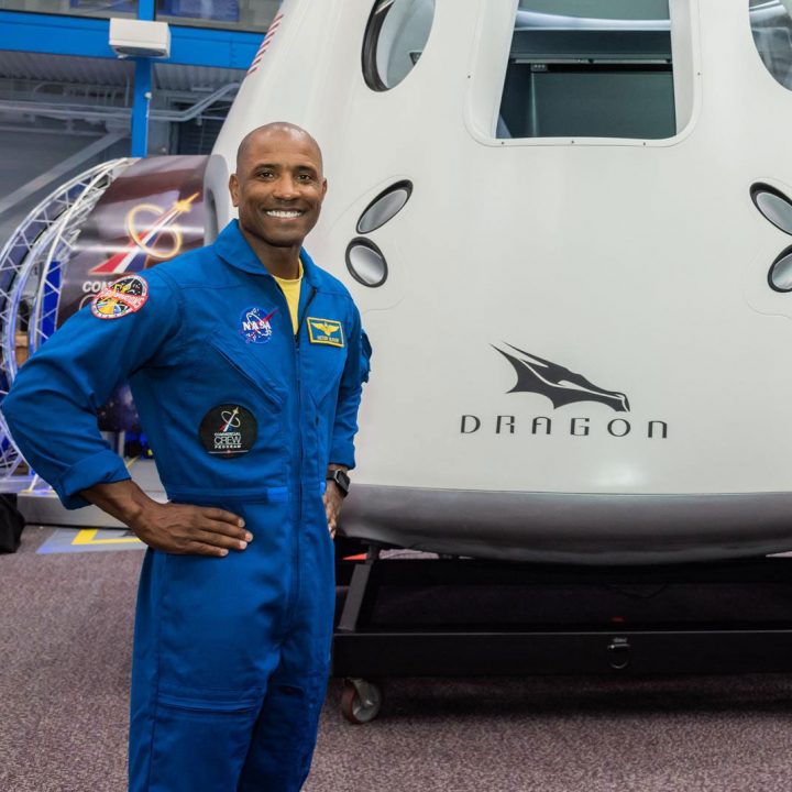 Astronaut Victor Glover stands in front of a SpaceX Crew Dragon capsule wearing a blue flight suit