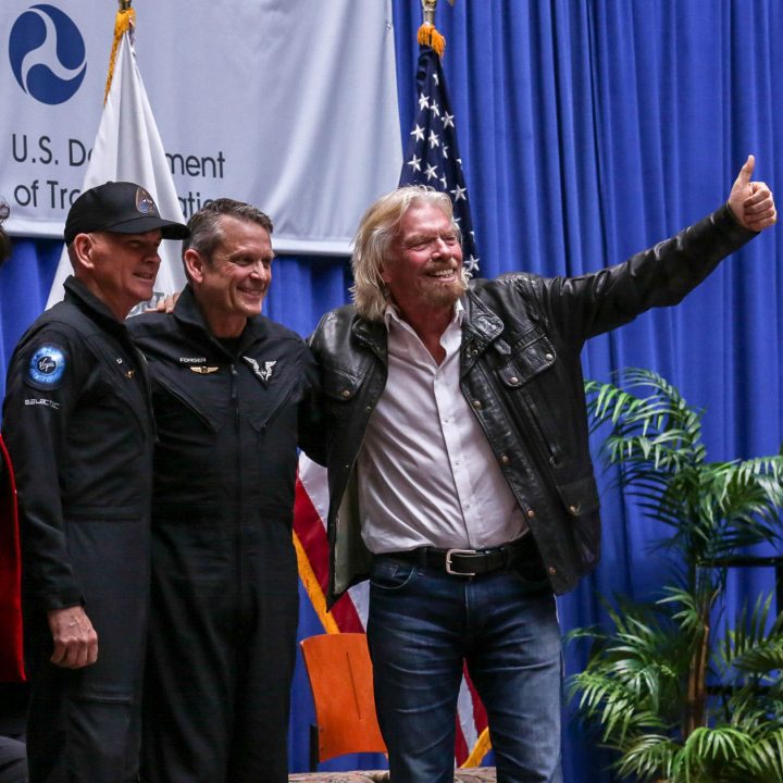 Two pilots in flight suits stand on a stage with Sir Richard Branson giving a thumbs up