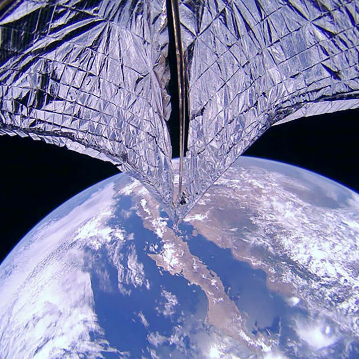 A silver mylar sail unfurls from a cubesat with Earth in the background