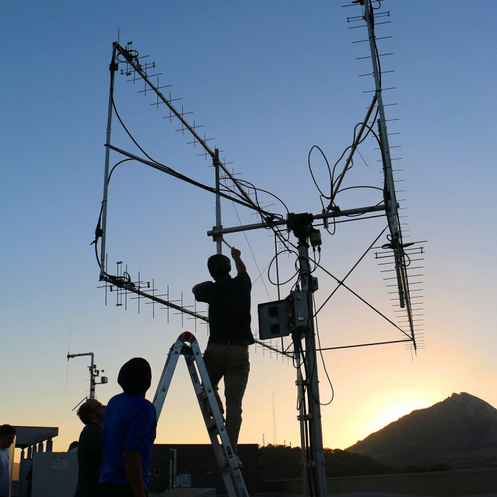 Two people fix an antenna on the roof of a building with the sun setting behind Bishop Peak in the background