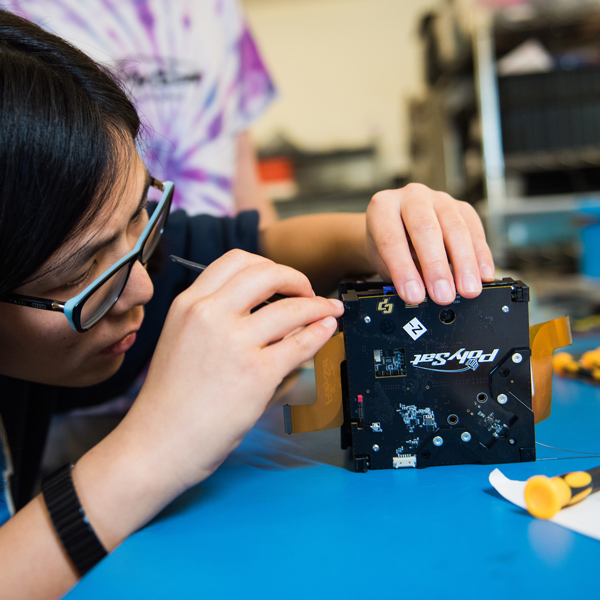 A student in glasses uses a tool on the shell of a small black cube satellite