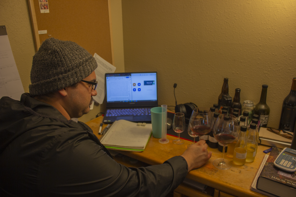 A student in a knit cap and glasses inspects wine samples while sitting at a desk in his house