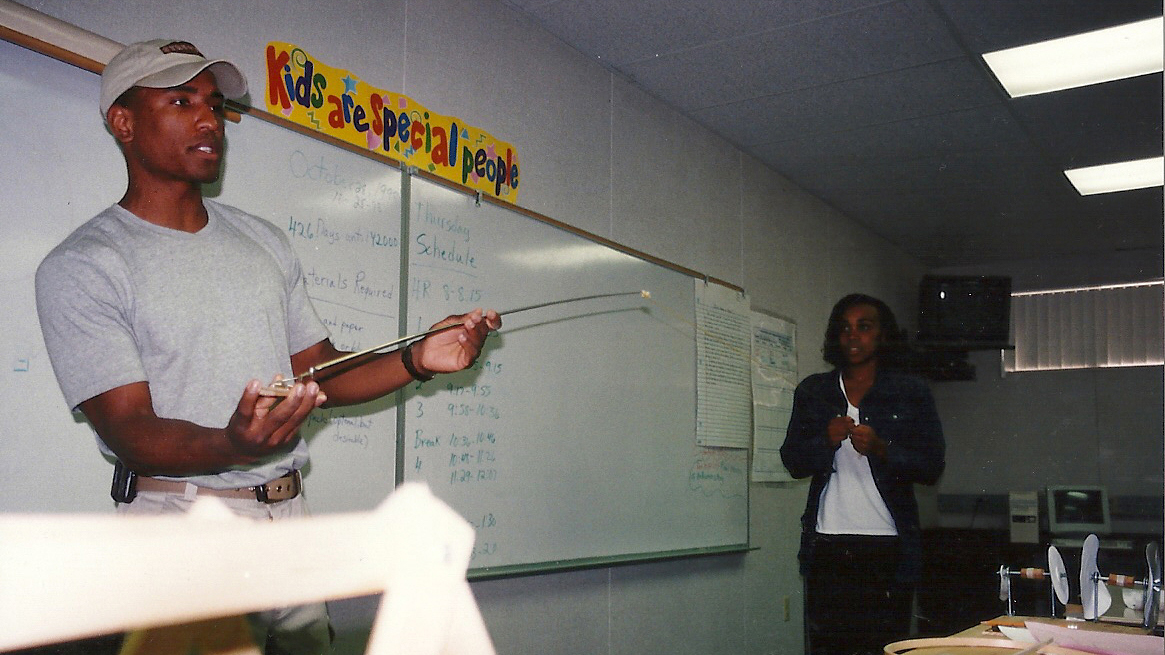 A younger Glover in a hat and t-shirt gestures in front of a middle school classroom whiteboard, while a young woman stands to the side watching.