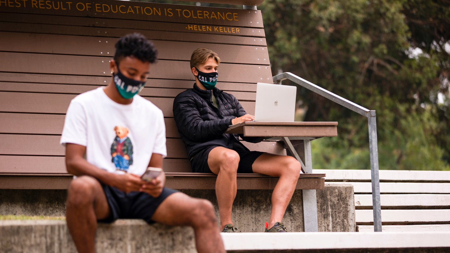 Two men wearing face masks sit near a bench