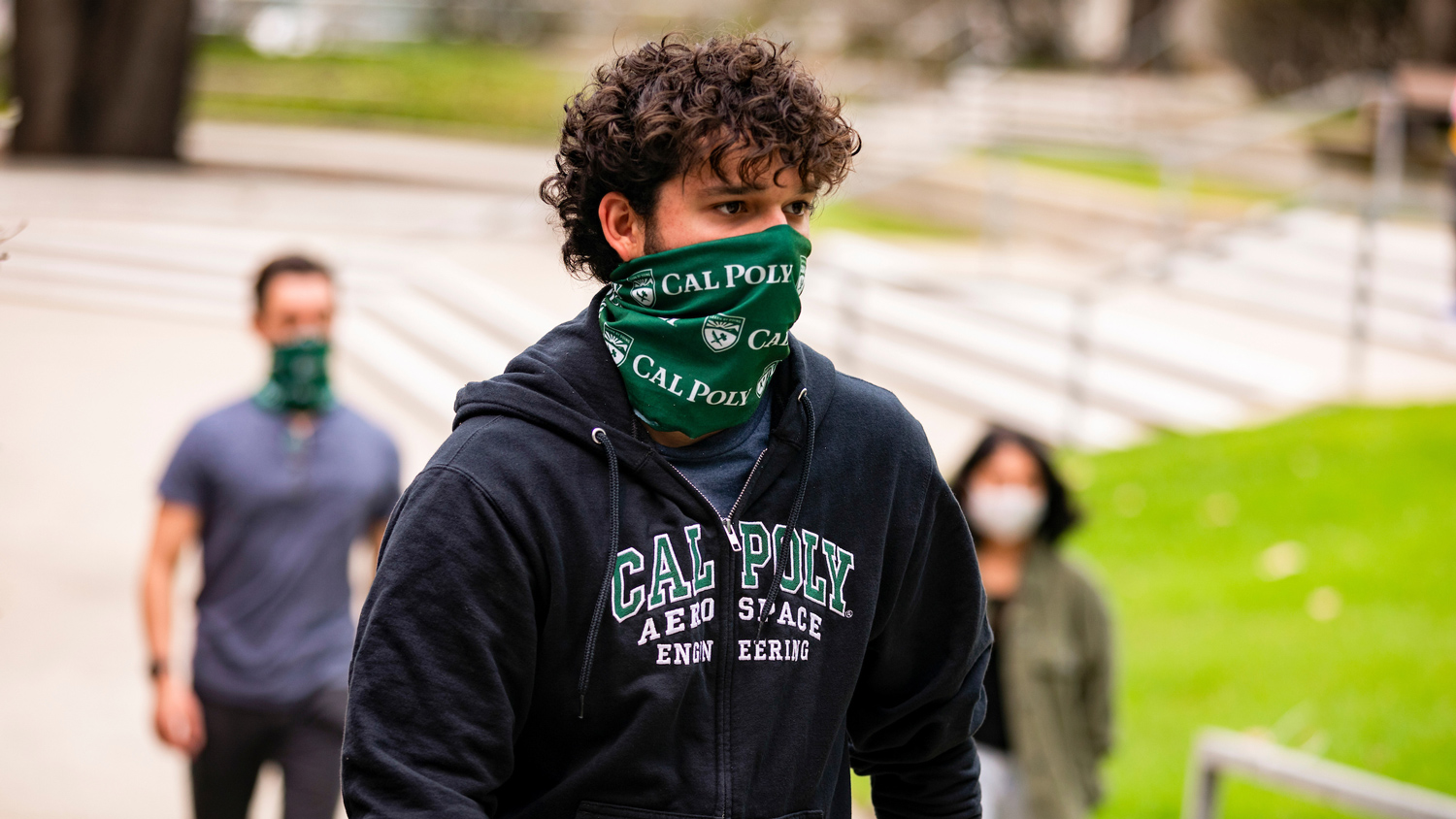 A man wearing a Cal Poly face covering and sweatshirt walks on campus with two people in masks behind him