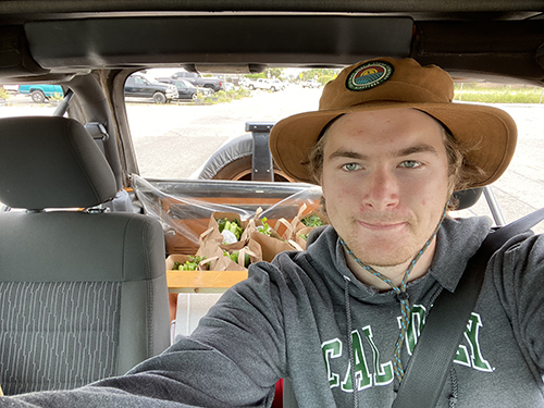 A selfie of a young man in a wide brimmed had and a Cal Poly sweatshirt in the front seat of a car filled with boxes of food.