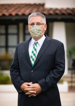 President Armstrong stands in front of the Cal Poly presidential residence wearing a dark suit and a facemask covered in CP marks