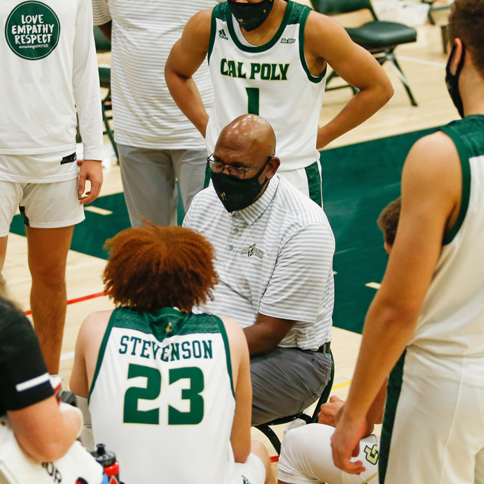A basketball coach wearing a mask sits surrounded by players