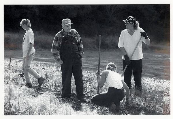 In a black and white photo, a man in overalls and a cap watches students working in a field.