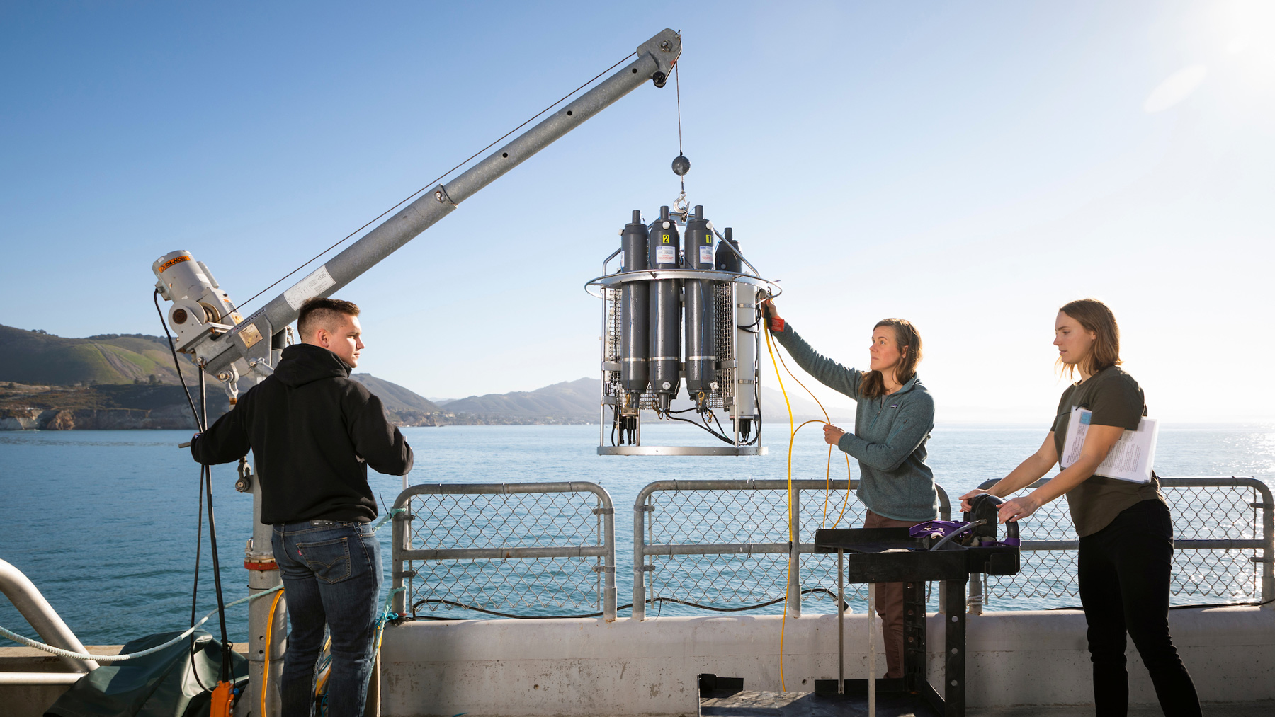 Three people lift machinery with a crane on the Cal Poly Pier